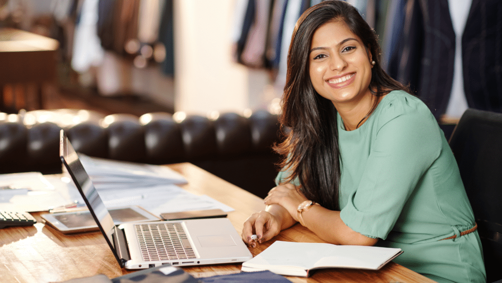 A business woman seated at her desk in her clothing shop looking for small business grants on her laptop.